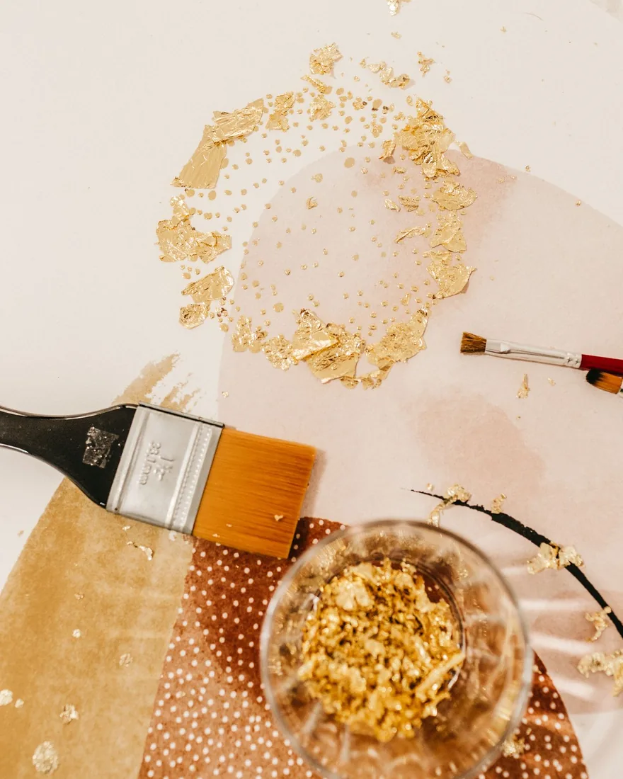 Pinceles planos, fragmentos de pan de oro y un recipiente de vidrio con restos dorados sobre una mesa de trabajo